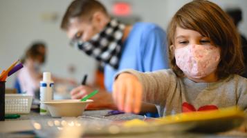 A child with shoulder length brown hair and a pink surgical mask reaches for some art supplies, while a man wearing a blue shirt and a black and white check bandana around his face draws in the background.