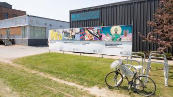 A photo of a square building with grey brown corrugated steel siding, and a sign that says BCA Studios: Burlington City Arts in blue lettering. A panel of 4 colorful murals, some grass and a bike rack with a bike in it sits in front of the building. A square blue grey building with a sign that says &quot;Maltex&quot; sits to the left.
