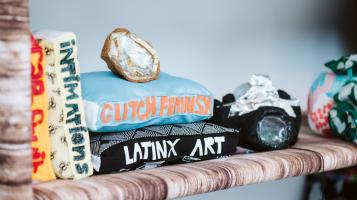 A photo of a detail of a soft sculpture, with books made out of textiles that say Intimations, Glitch Feminism, and Latinx Art. They sit on a shelf made from brown and white variegated fabric alongside a black camera made of fabric and a silver and grey geode made of fabric.
