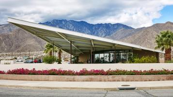 A photograph of a modern building with. white triangular dramatically slanted roof that overhangs a small glass building with brown and blue mountains and desert in the background.