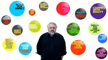 a light skinned man with grey curly hair and a bread wearing a black shirt against a white backdrop with large brightly colored buttons with text arranged around him