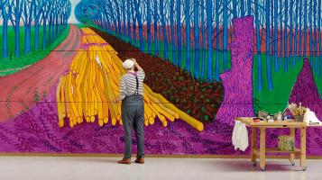 A photograph of David Hockney, a light skinned man with white hair wearing grey trousers and suspenders, working on a large painting with abstract shapes in fushia, green, saffron yellow, and teal.