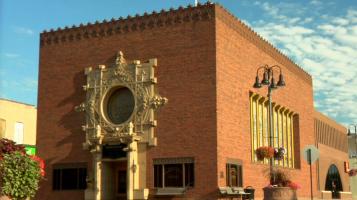 A square, red brick building with crenellated trim around the flat roof and ornate cream colored stone detailing around a circular window and doorway, with a blue sky with rippling clouds in the background