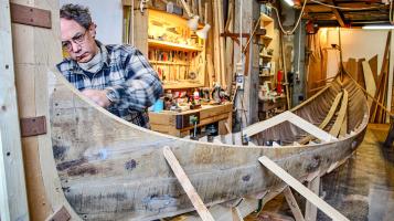 A photograph of a light skinned man wearing a blue and white plaid shirt working on building a grey gondola that is propped up with wooden stakes. wooden shelves filled with tools and wires hang down in the background of the workshop.