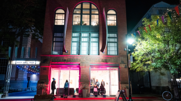 A photo of the BCA Center at night. A narrow 4 story, red brick building with tall, arched windows, and aqua vertical banners, lit up inside, with people visible through the gallery doors