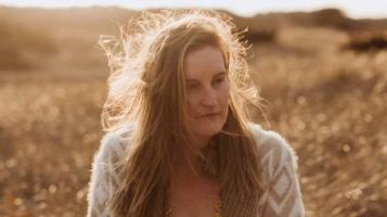 a light skinned woman with red hair seen from the chest up stands in a field that appears golden, backlit by sun