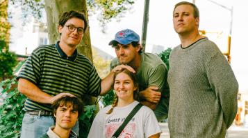 Four light skinned men and one woman are clustered together. One man wearing round glasses and green and black striped polo shirt stands on the left, with his hands on top of the head of a man who is sitting on the ground and the seated woman. Another man wearing a blue baseball cap leans over the seated woman and stand next to another man wearing a light green sweatshirt.