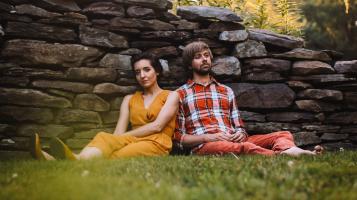 A man and a woman sit on the ground, leaning together against a stone wall. The woman has light skin and dark hair and wears a dark yellow  dress and shoes.The man has light skin and light brown hair and wears a red white and blue plaid shirt and red pants