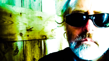 A close up of the face of a light skinned man with a silver beard and hair, wearing dark sunglasses against a barn board background