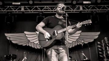 A black and white photograph of a light skinned man with a dark beard and hair playing a guitar and singing into a microphone on a stage, framed by a decorative pair of wooden wings