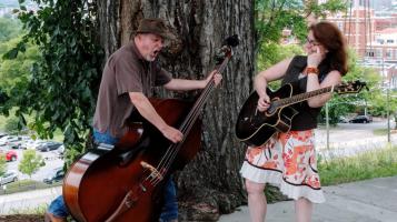 A light skinned man wears a brown shirt and cowboy hat and plays a stand up bass, tilting it as he sings. A light skinned woman with brown hair and a floral print skirt plays a guitar and laughs. They stand in front of a large tree, with brick building s and a parking lot visible in the background