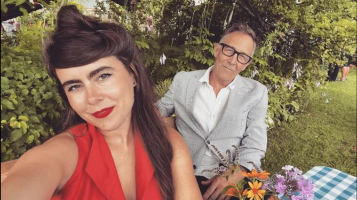 A light skinned young woman with brown hair and a red dress sits on a picnic blanket next to an older light skinned man with silver hair wearing a grey suit