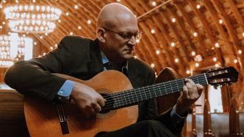 A light skinned bald man wearing glasses and a dark suit plays an acoustic guitar. He sits in a pew under a wooden vaulted ceiling studded with points of light