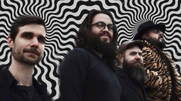 Four men, all with light skin, dark hair, and beards stand against a backdrop of wavy black and white starbursts