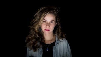 A woman with light skin and dark red hair is seen from the chest up, against a chiaroscuro-like black background