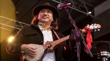 A tibetan man seen from the waist up stands on stage at a microphone as he plays at dramyin, a traditional Himalayan lute