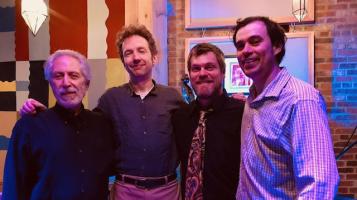 Four light skinned men, seen from the waist up stand with their arms around each other's shoulders in front of a brick wall painted in blocks of red, yellow, and white.