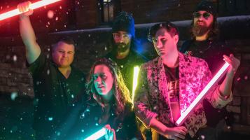 Four light skinned men and one light skinned woman stand in the snow at night, illuminated by the colorful fluorescent tube lights they hold