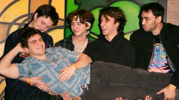 three young men with light skin tones and one with a medium skin tone laugh as they all hold another light skinned young man, laying horizontally