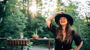 A light skinned woman with brown hair is seen from the waist up, as she laughs and holds a brown wide brimmed hat on her head. Potted plants and green trees are out of focus behind her