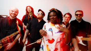 A dark skinned woman wearing a white dress patterned with colorful birds stand in front of five light skinned men wearing black. One man holds a saxophone, one drum sticks, and one a guitar. The image is slightly warped from a time lapse with red streaks