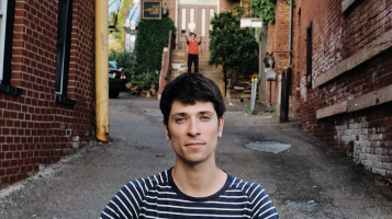 A light skinned man with dark hair and a black and white striped tee shirt is seen from the shoulders up. He stands in an alleyway, between brick buildings and in the background another man is positioned to look as is he is standing on the first man's head.