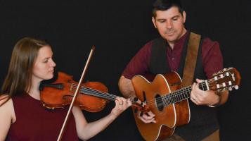 Against a black backdrop, a light skinned woman, seen from the chest up plays the violin, and a light skinned man seen from the waist up plays a guitar