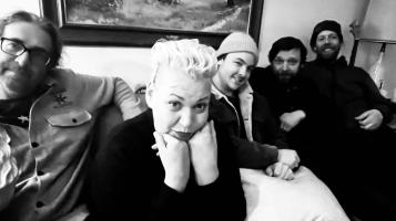 A black and white image of four light skinned men and one light skinned woman sitting on a couch. The woman who has a bleached blond pompadour leans forward and rests her chin on her hands