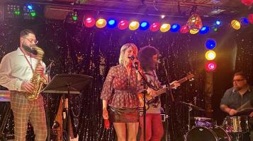 A four person band plays on a stage with a glittery backdrop and a row of red, yellow, and blue lights. A light skinned woman wearing a sparkly miniskirt sings into a mic in the center of the stage. On the left a light skinned man plays the saxophone, and on the right a light skinned man plays bass, and another sits behind a drum set