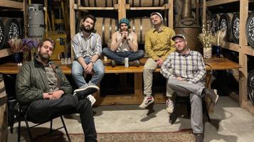 Five light skinned men sit on a table and chairs in a distillery, with barrels of whiskey stacked on shelves behind them and a maroon oriental rug at their feet