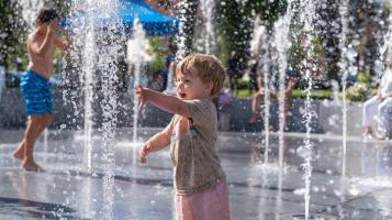 A light skinned young boy reaches out to touch one of the streams of water jetting up from the ground around him. Another boy runs across the splash fountain in the background