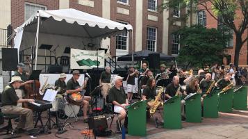 An 18 piece jazz band plays outdoors on the brick surface of the Church Street Marketplace, with a white tent over a raised stage and Burlington's City Hall, a brick and white stone building behind them.