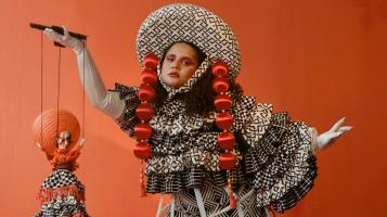 A medium skinned woman with dark hair wears an elaborate black and white ruffled patterned ruffled dress with a matching wide brimmed hat trimmed with two long strands of red balls that hang down beside her face. She stands against an orange backdrop and hold a marionette dressed in a similar style in one hand