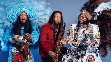 Three dark skinned men play musical instruments against a light blue background. On the right, one man wears a large blue feathered headdress, a blue patterned and ruffled coat, and plays the tambourine and sings.  In the middle, one man wears a red blazer and plays the saxophone. On the left one man has orange and yellow stripes painted across his cheeks and wears a large brown, black, and white feathered headdress and a white embroidered and patterned coat  while he plays the tambourine