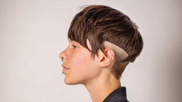 A profile headshot of a non binary person with a medium skin tone, short brown asymmetrical hair with a shaved undercut, and a nose ring
