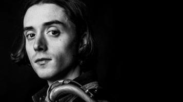 A black and white profile headshot of a light skinned young man with a saxophone neck hooked over his shoulder
