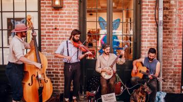 Four light skinned men play musical instruments in front of a brick building. One wears a wide brimmed hat and plays a stand up bass, one wears a white shirt and black suspenders and plays the fiddle, one sits and plays the banjo, and one sits and plays an acoustic guitar