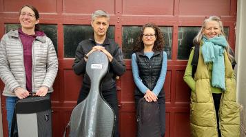 Four people stand against a red garage door. Three are light skinned women and one is a light skinned man holding a grey cello case