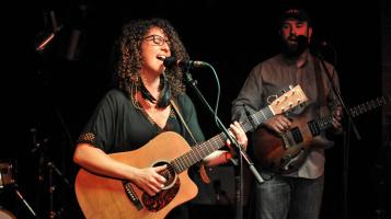 A light skinned woman with curly dark hair and dark rimmed glasses plays an acoustic guitar and sings into a microphone.