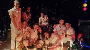 Nine people sit, crouch, and stand clustered together. They all wear off white sleeveless jumpsuits. Some hold gold streamers. A sparkly curtain and stage lights are in the background