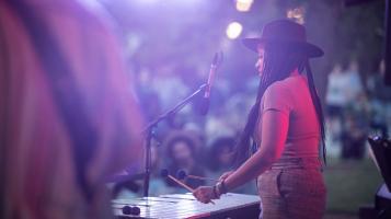 A dark skinned woman with long black braids wears a brown fedora and plays the vibraphone at an outdoor concert. She's seen from the side through a hazy, purple twilight and people sitting on the grass are visible in the background.