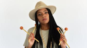 A dark skinned woman with long black braids wears a white fedora and top and holds four vibraphone sticks