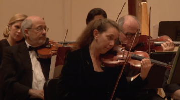 A group of people sit together, all playing violins. Two are light skinned men, both with balding, grey hair. One is a light skinned woman with brown hair, and one partially obscured, is a light skinned woman with blonde hair