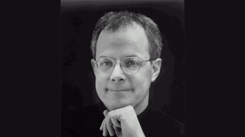 A head shot of a light skinned man wearing round wire rimmed glasses and resting his chin on his hand against a black backdrop