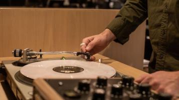 A cream colored record spins on an elaborate turntable set up, and the hands of a light skinned man wearing an olive green shirt can be seen lifting the needle. The back of a blonde wood banquette blond wood piece is in the background