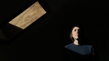 A light skinned man with light brown hair seen from the shoulders up, with his face half obscured by a dark background, interrupted only by a geometric wooden form floating above him