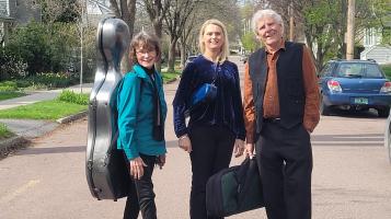 An older light skinned man and two light skinned women stand in the middle of a street. One woman wears a cello case on her back, one wears a rectangular flute case on a strap across her body, and the man holds a violin case