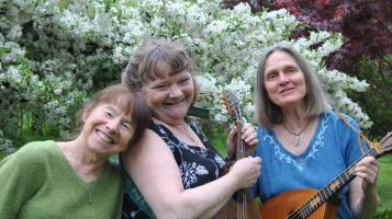 Three light skinned women with greying brown hair stand together in front of a flowering white cherry blossom tree. One woman holds a mandolin, one a guitar, and the third rests her head on the shoulder of the second