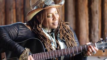 A dark skinned woman with long brown braids is seen from the waist up, seated. She wears a straw cowboy hat and plays the guitar