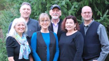 Three light skinned older men stand behind three light skinned older women, with leafy trees in the background